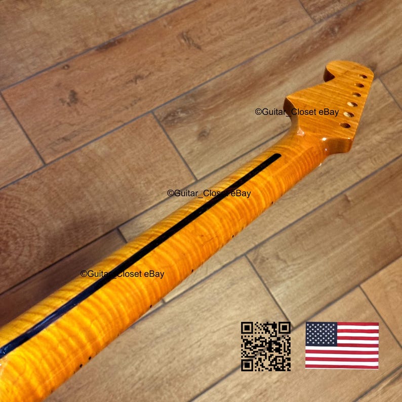 May include: A close-up of a guitar neck with a flamed maple finish. The neck has a dark blue stripe down the center and is angled towards the top of the image. The headstock has tuning peg holes. The background is a wooden floor.