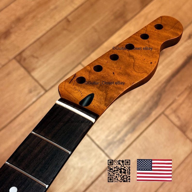 May include: A guitar neck with a dark fretboard and a light brown headstock. The headstock has six tuning peg holes and a dark, oval-shaped truss rod access. The wood grain is visible. The image includes a QR code and the American flag.