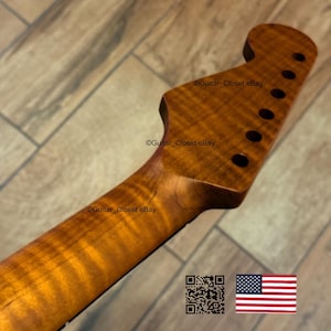 May include: A close-up of a guitar neck made of figured wood, featuring a headstock with tuning peg holes. The wood grain has a warm, amber hue. The image includes the text "Guitar_Closet eBay" and a US flag.