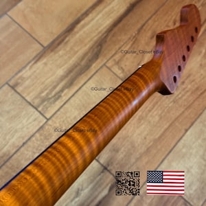 May include: A close-up of a guitar neck made of wood with a rich, amber color and a prominent grain pattern. The headstock features tuning peg holes. The text "Guitar_Closet eBay" is visible. An American flag is in the bottom right corner.