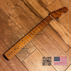 May include: A light brown maple guitar neck featuring a prominent flame grain pattern, a smooth fretboard with dark circular position markers, and a headstock with six pre-drilled tuning peg holes. The neck displays a natural wood finish.