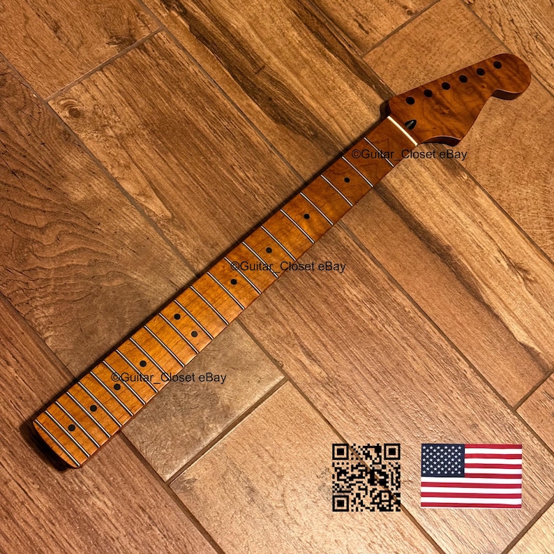 May include: A light brown maple guitar neck featuring a prominent flame grain pattern, a smooth fretboard with dark circular position markers, and a headstock with six pre-drilled tuning peg holes. The neck displays a natural wood finish.