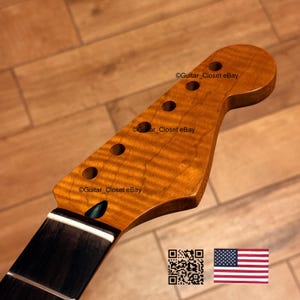 May include: A guitar neck with a flamed maple headstock and a dark fretboard. The headstock has six tuning peg holes and a natural finish. The neck is angled and has a white stripe at the base. The American flag is in the bottom right corner.
