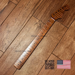 May include: A guitar neck featuring a flamed maple wood grain pattern with a natural brown finish. The neck includes a fretboard with dark circular fret markers and silver frets. The headstock has six pre-drilled holes for tuning pegs.