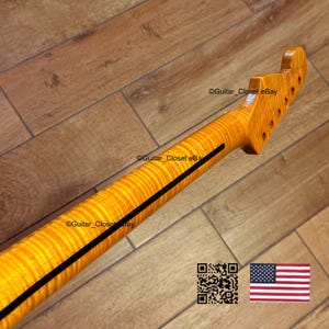 May include: A close-up of a guitar neck with a flamed maple finish. The neck is a light amber color with a dark black stripe down the center. The headstock has tuning peg holes. The background is a wooden floor.