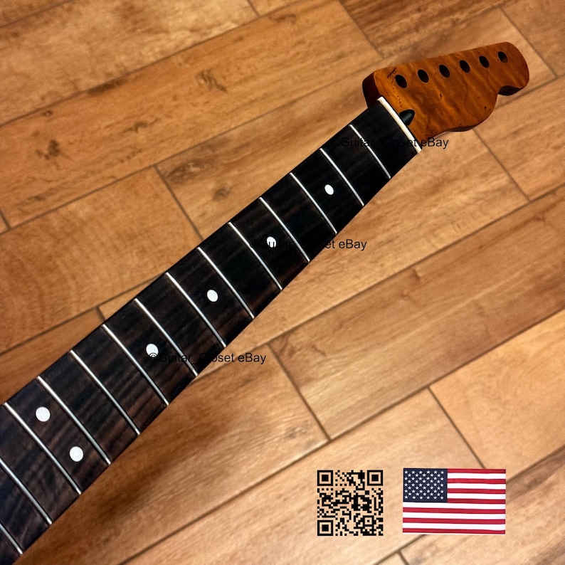 May include: A guitar neck with a dark fretboard and white fret markers. The headstock is a lighter wood with tuning peg holes. The neck is angled on a wooden surface. The American flag and a QR code are in the bottom right corner.