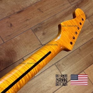 May include: A close-up of a guitar neck with a flamed maple finish. The neck has a dark stripe down the center and six tuning peg holes. The headstock is shaped with a rounded top. The wood grain is visible.