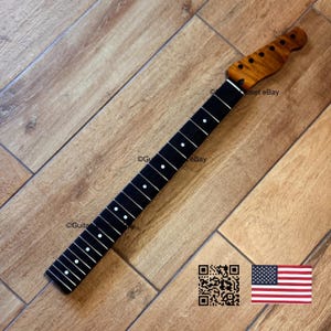 May include: A guitar neck with a dark fretboard and white dot inlays. The headstock is a lighter wood with a flamed grain pattern. The neck is angled on a wooden surface. A QR code and American flag are in the lower right corner.