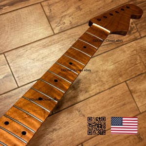 Puede incluir: Un m&aacute;stil de guitarra con un acabado de veta de madera en tonos &aacute;mbar c&aacute;lidos, con un clavijero prominente, trastes plateados y marcadores de traste circulares oscuros. La superficie muestra un patr&oacute;n de madera natural en toda su longitud.