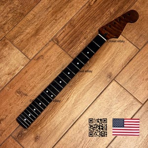 May include: A guitar neck with a dark brown headstock and a black fretboard with white dot inlays. The neck is angled on a wooden surface. The image includes a QR code and an American flag.