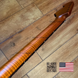 May include: A close-up of a guitar neck made of figured maple wood. The neck has a warm, amber color with a prominent wood grain pattern. The fretboard is dark, and the headstock is visible. The image includes a QR code and the American flag.