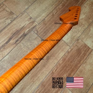 May include: A guitar neck with a flamed maple finish. The neck is a light amber color with a prominent wood grain pattern. The headstock has pre-drilled holes for tuning pegs. The image includes a QR code and the American flag.