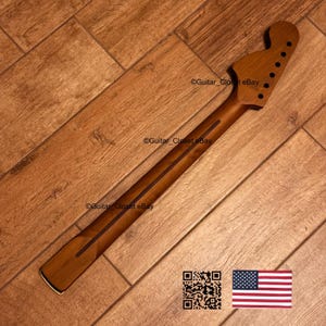 May include: A wooden guitar neck with a headstock featuring six tuning peg holes. The neck has a dark brown stripe down the center and is set against a wooden floor. The image includes a QR code and an American flag.