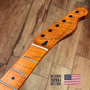 May include: A close-up of a guitar neck and headstock made of light brown wood. The headstock has six tuning peg holes and a dark blue inlay. The fretboard has white frets and a dark blue dot inlay. The wood grain is visible.