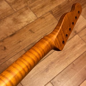 May include: Close-up of a guitar neck with a natural wood finish. The neck has a light brown color with a prominent wood grain pattern. The headstock features six tuning peg holes.