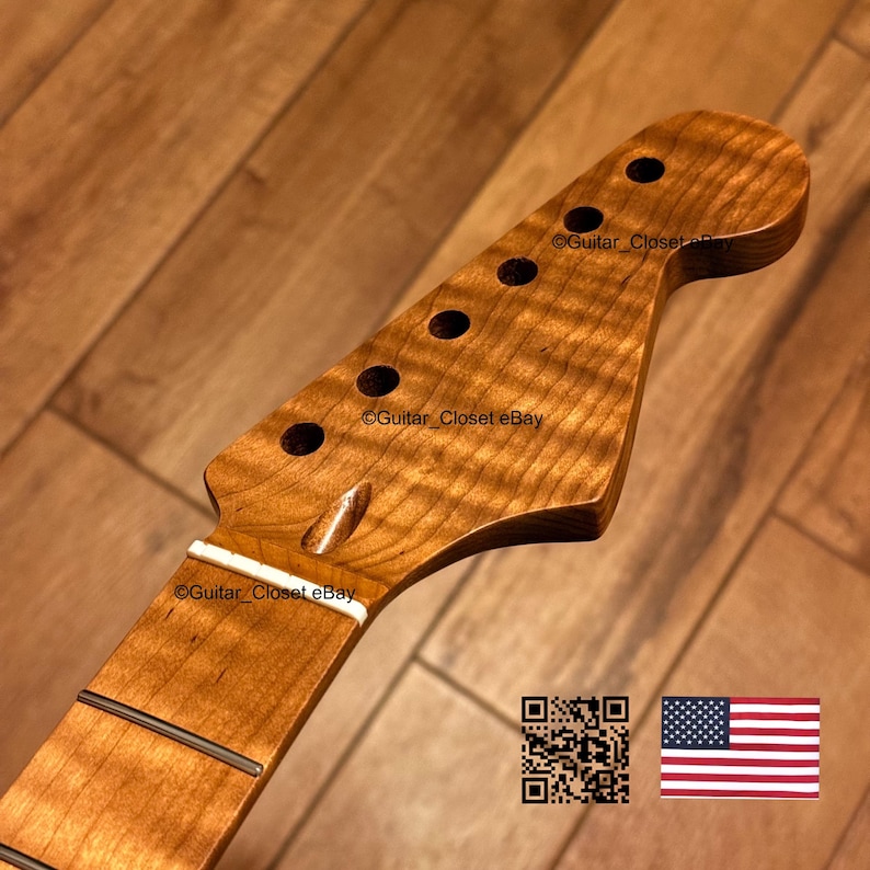 K&ouml;nnte beinhalten: Ein Gitarrenhals mit einer Kopfplatte aus geflammtem Ahorn in nat&uuml;rlichem Bernsteinton, sechs Mechanikbohrungen, einem wei&szlig;en Sattel und einem Griffbrett mit sichtbaren Metallb&uuml;nden. Die Holzmaserung zeigt ein deutliches Wellenmuster.