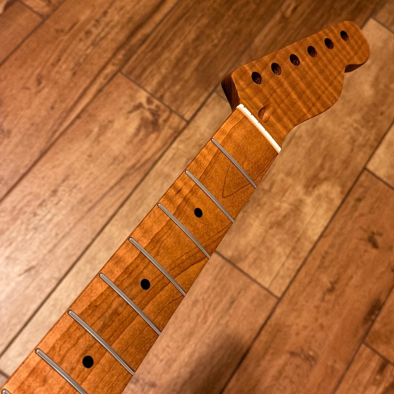 Op de afbeelding: Een gitaarhals gemaakt van amberkleurig gevlamd esdoornhout, voorzien van een kop met zes gaten voor stemmechanieken, een witte topkam en een toets met zilverkleurige metalen frets en donkere ronde positiemarkeringen langs de hals.
