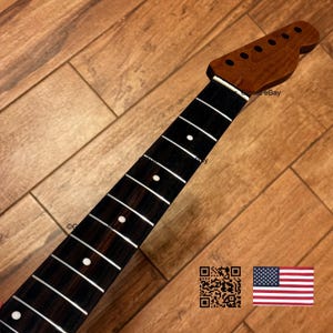 May include: A guitar neck featuring a dark brown wooden headstock with six tuning peg holes and a dark, grained fretboard. The fretboard includes silver metal frets and white circular position markers. The neck is shown at an angle.