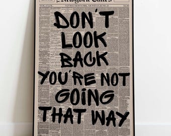 May include: A framed print featuring a vintage newspaper background with bold black text that reads "DON'T LOOK BACK YOU'RE NOT GOING THAT WAY." The print is from The New York Times, dated June 21.
