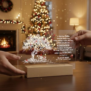 May include: A clear acrylic memorial plaque with a tree design, illuminated by lights. The tree features red birds and the text "A limb has fallen...Grieve not for me...Remember...while I was STRONG. ELIJAH REID July 12, 1968 - August 5, 2024."