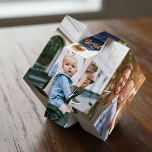 May include: A personalized photo cube featuring multiple family photos. The cube is made of white blocks and displays various images of a baby, a man, and a couple. The cube is displayed on a wooden surface.