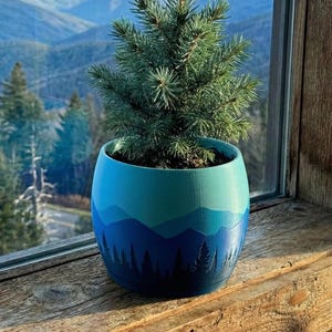 May include: A small evergreen tree in a blue and teal painted pot with a mountain and tree silhouette design. The pot is sitting on a wooden windowsill with a scenic mountain view in the background.