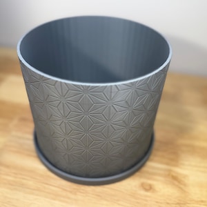Puede incluir: Una maceta cil&iacute;ndrica gris con un patr&oacute;n geom&eacute;trico de estrellas. La maceta tiene un platillo a juego y est&aacute; hecha de un material s&oacute;lido. El dise&ntilde;o est&aacute; en relieve en el exterior, agregando textura e inter&eacute;s visual a la pieza.