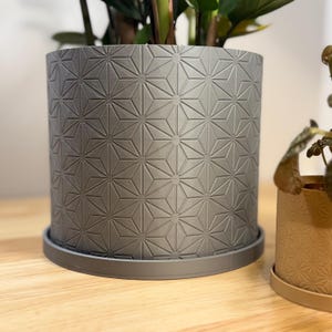 Puede incluir: Una maceta cil&iacute;ndrica gris con un patr&oacute;n geom&eacute;trico de estrellas. La maceta se asienta sobre un platillo a juego. La maceta est&aacute; llena de plantas verdes. En el fondo se ve una maceta m&aacute;s peque&ntilde;a de color beige.