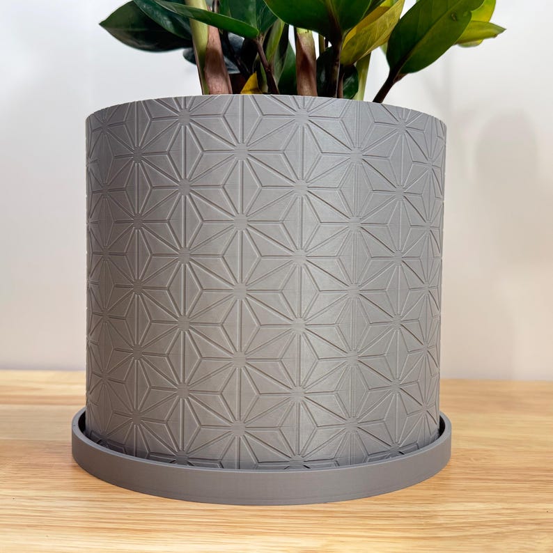 Modern Matte Ash Gray 3D printed indoor planter with geometric star (Hexstar) pattern texture, cylindrical shape, and matching drip tray, shown with a green houseplant on a wooden surface.