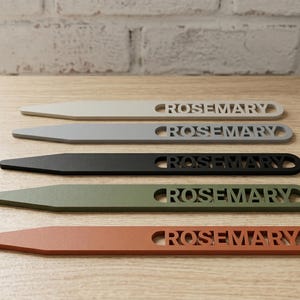 May include: Five wooden plant markers in a row, each with the word "ROSEMARY" cut out. The markers are in shades of beige, gray, black, olive green, and orange. They are arranged on a light wood surface.