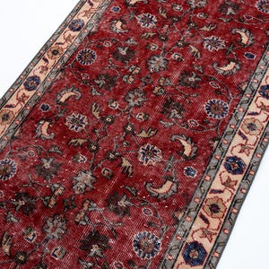 May include: A long, rectangular runner rug featuring a deep red central field adorned with intricate floral and vine motifs in shades of blue, beige, and dark gray. The rug is framed by a decorative border with repeating geometric patterns.