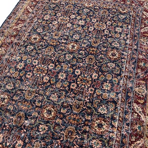 May include: A rectangular area rug featuring a dark navy blue central field densely patterned with intricate floral and botanical motifs in shades of red, cream, and gold, framed by a wide, ornate border in deep red with repeating geometric designs.