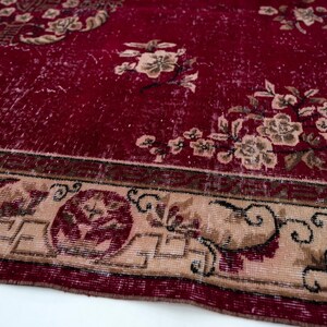 May include: Close-up of a distressed red rug with floral patterns. The rug features a faded appearance with beige and brown floral designs scattered across the red background. A beige border with intricate patterns frames the rug.