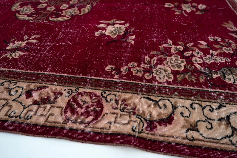 May include: Close-up of a distressed red rug with floral patterns. The rug features a faded appearance with beige and brown floral designs scattered across the red background. A beige border with intricate patterns frames the rug.