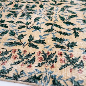 May include: Close-up of a beige rug with a floral pattern. The rug features green and blue leaves with red flowers along the bottom edge. The rug has a distressed, vintage look.