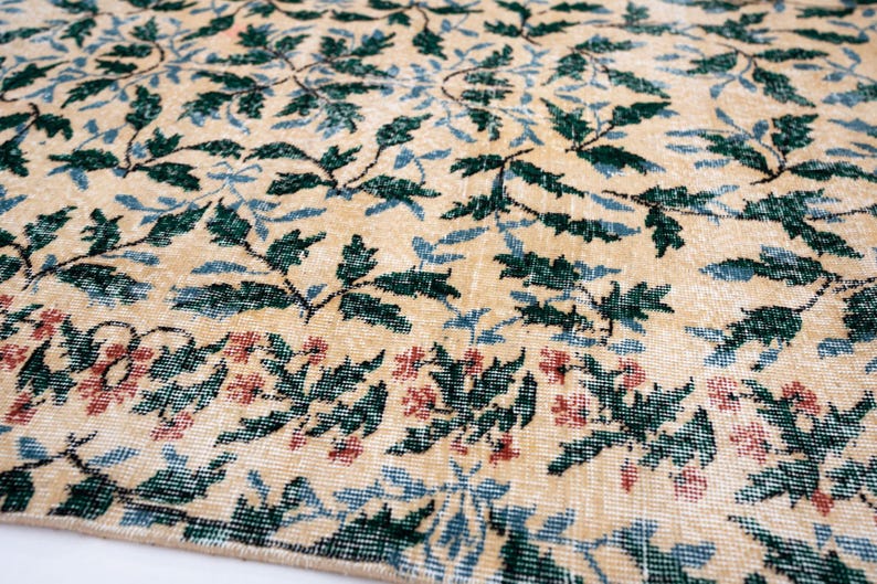 May include: Close-up of a beige rug with a floral pattern. The rug features green and blue leaves with red flowers along the bottom edge. The rug has a distressed, vintage look.