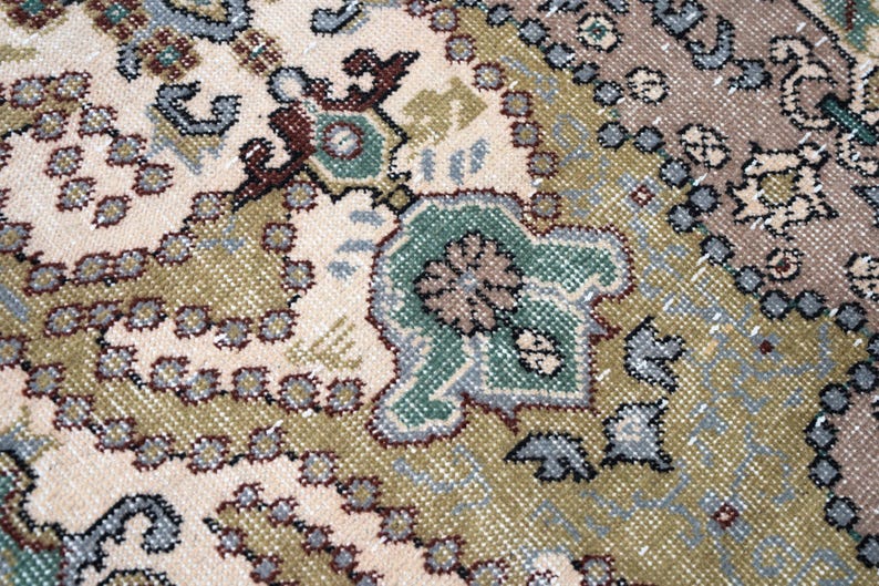 May include: A woven rug featuring a complex pattern with a cream-colored background, olive green sections, and accents in muted blue, brown, and dark gray. The design includes repeating floral motifs, geometric shapes, and intricate border details.