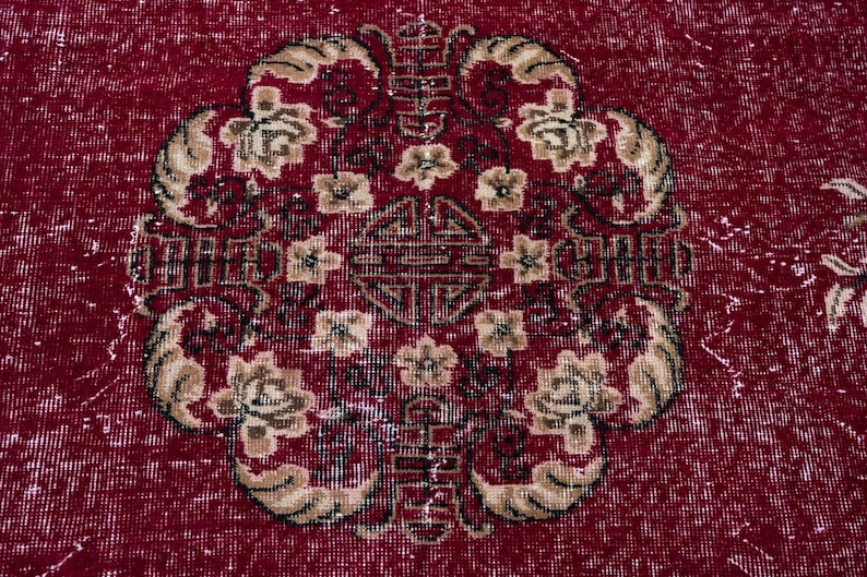 May include: A close-up of a red rug with an ornate floral design. The central motif features a symmetrical arrangement of flowers and geometric shapes in beige and dark brown. The rug has a distressed, vintage appearance.