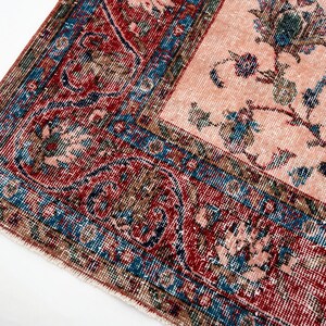 Pu&ograve; includere: Primo piano di un tappeto vintage dall'aspetto usurato e sbiadito. Il tappeto presenta un campo centrale rosa pallido con motivi floreali blu e verdi. Il bordo &egrave; un mix di rosso, blu e marrone, con disegni intricati.