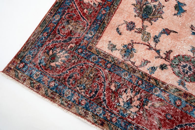 Pu&ograve; includere: Primo piano di un tappeto vintage dall'aspetto usurato e sbiadito. Il tappeto presenta un campo centrale rosa pallido con motivi floreali blu e verdi. Il bordo &egrave; un mix di rosso, blu e marrone, con disegni intricati.