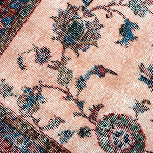 Pu&ograve; includere: Primo piano di un tappeto vintage con un motivo floreale sbiadito. Il tappeto presenta uno sfondo color pesca chiaro con dettagliati disegni floreali e di vite nelle tonalit&agrave; del blu, verde e bordeaux. Il bordo &egrave; un mix di rosso, blu e verde.
