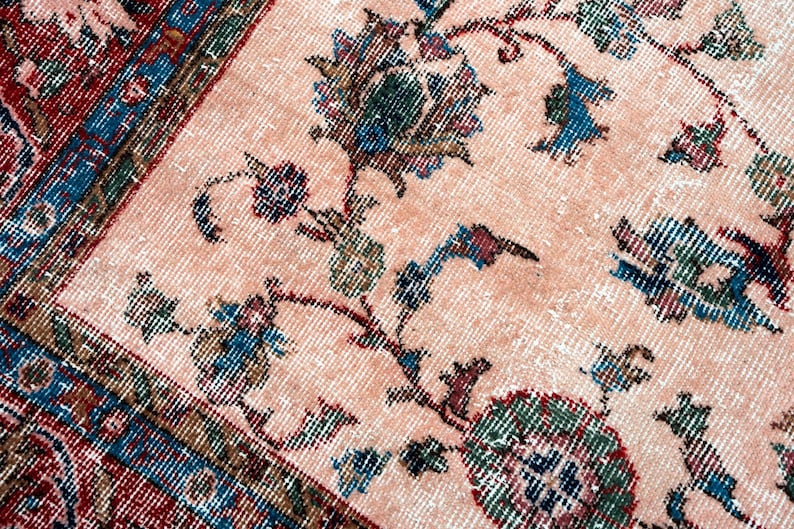 Pu&ograve; includere: Primo piano di un tappeto vintage con un motivo floreale sbiadito. Il tappeto presenta uno sfondo color pesca chiaro con dettagliati disegni floreali e di vite nelle tonalit&agrave; del blu, verde e bordeaux. Il bordo &egrave; un mix di rosso, blu e verde.