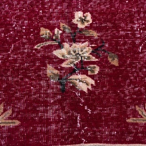 May include: Close-up of a red rug with a floral pattern. The rug has a distressed look with white accents. The floral design features beige flowers and leaves with black stems, creating a vintage aesthetic.