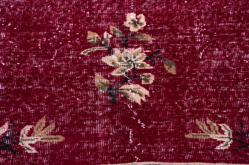 May include: Close-up of a red rug with a floral pattern. The rug has a distressed look with white accents. The floral design features beige flowers and leaves with black stems, creating a vintage aesthetic.