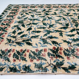 May include: Beige area rug with a floral pattern. The rug features a central design of green leaves and blue accents, framed by a border of red and green floral motifs. The rug has a distressed, vintage appearance.