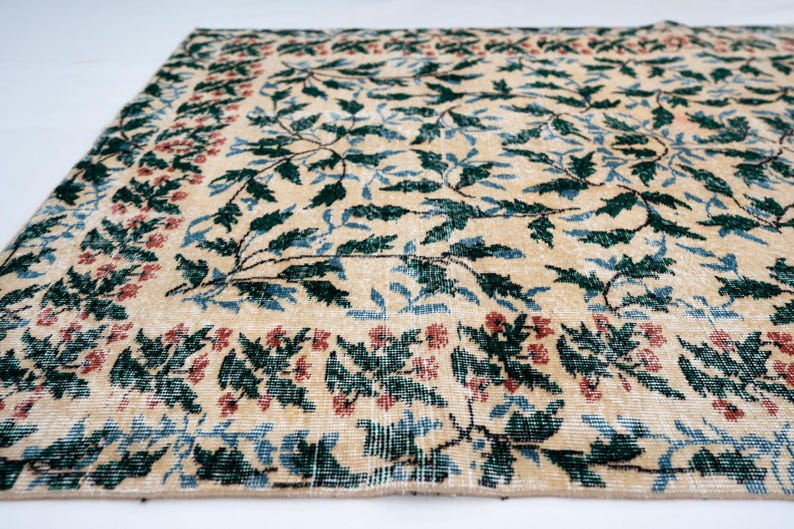 May include: Beige area rug with a floral pattern. The rug features a central design of green leaves and blue accents, framed by a border of red and green floral motifs. The rug has a distressed, vintage appearance.