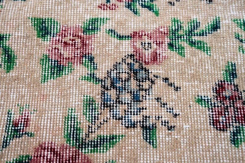 Pode incluir: Close-up de um tapete bege com um padr&atilde;o floral. O tapete apresenta desenhos repetidos de flores cor-de-rosa com folhas e caules verdes. Um elemento de design central tem detalhes em azul e preto.