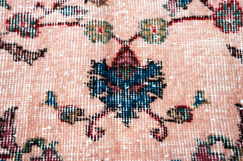 Pu&ograve; includere: Primo piano di un tappeto vintage con un motivo floreale dettagliato. Il tappeto presenta uno sfondo color pesca chiaro con disegni intricati in verde acqua, bordeaux e verde oliva. Il motivo centrale &egrave; una composizione floreale stilizzata.