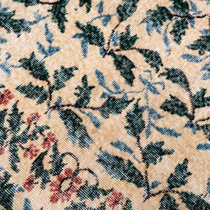 May include: Close-up of a beige rug with a floral pattern. The rug features green leaves, blue accents, and red flowers. The design is intricate and detailed, with a vintage or antique aesthetic.