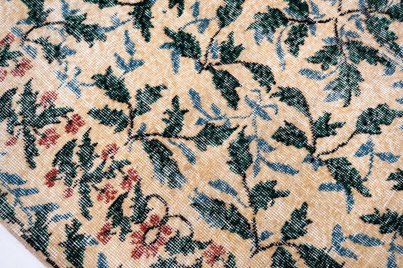 May include: Close-up of a beige rug with a floral pattern. The rug features green leaves, blue accents, and red flowers. The design is intricate and detailed, with a vintage or antique aesthetic.